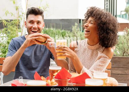 Felice affamata coppia multiculturale gustare hamburger e birre fredde in un ristorante all'aperto o un pub ridendo felice a una buona scherzata Foto Stock