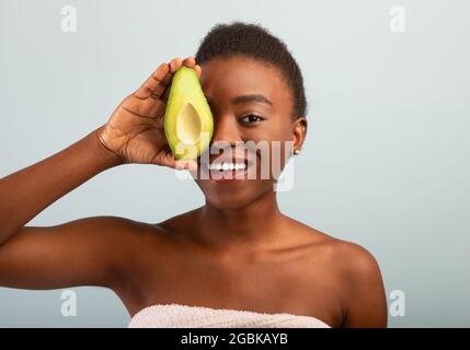 Felice african american giovane donna che tiene fresco avocado metà e coprente occhio, sorridente a macchina fotografica sopra sfondo grigio Foto Stock
