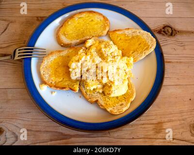 Brunch o pranzo anticipato su un tavolo all'aperto con uova strapazzate su pane tostato bianco su un piatto bianco con bordi blu su un tavolo di legno Foto Stock