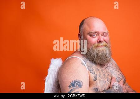 Il grasso uomo doubter con barba, tatuaggi e ali agisce come un angelo Foto Stock