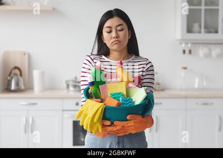 Donna asiatica esausta con attrezzi di pulizia in piedi in cucina Foto Stock