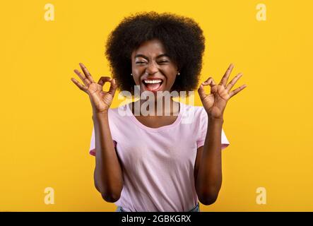 Tutto va bene. Giocosa donna nera sopravegata facendo gesto di approvazione, sorridente, isolata su sfondo giallo Foto Stock