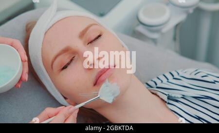 Mani Cosmetician applicazione maschera crema sul viso della giovane donna presso il salone di bellezza spa. Trattamenti per la cura della pelle del viso. Foto Stock