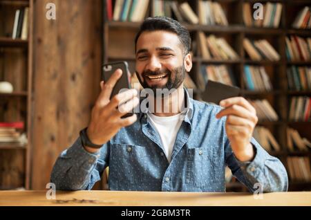 Felice gioioso giovane indiano che paga online con carta di credito, utilizzando l'app per smartphone per lo shopping online, ragazzo orientale misto-corsa con carta di debito e telefono cellulare, il trasferimento di denaro Foto Stock