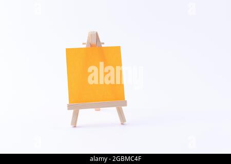 Easel in legno in miniatura con tela quadrata colorata vuota o carta Memo - Mockup. Mini supporto in legno con tavola da disegno pulita su sfondo bianco, spazio per la copia Foto Stock