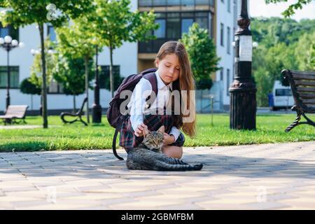 Ragazza piccola carina in uniforme e con zaino che accarezza un gatto vagante sulla preda Foto Stock