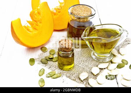 Olio di zucca in una fiala di vetro, salsa barca e un vaso su tela, semi sul tavolo, fette di verdure su sfondo di legno Foto Stock