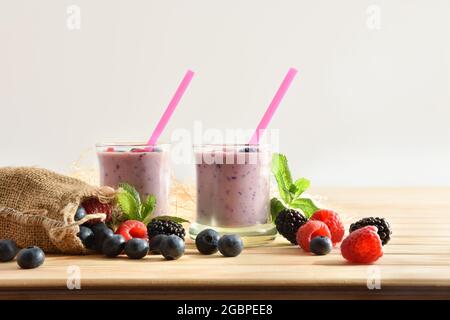 Frullato di yogurt alla bacca di bosco in bicchieri di vetro su tavolo di legno con sacco pieno di mirtilli, more lamponi bianco sfondo isolato Foto Stock