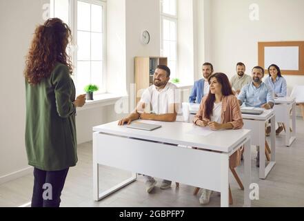 Il Business Coach parla con studenti adulti felici che si trovano alle scrivanie in classe Foto Stock