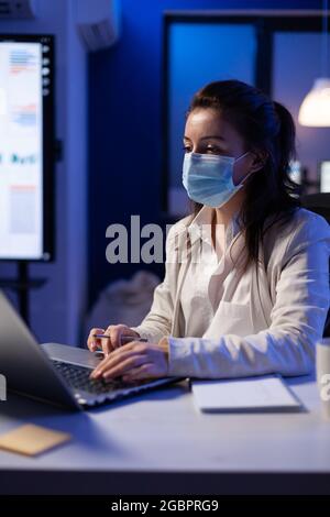 Manager donna con maschera facciale che lavora straordinario in un nuovo ufficio aziendale normale che guarda al progetto di team su un notebook professionale. Analizzare i documenti finanziari seduti alla scrivania durante la pandemia globale Foto Stock