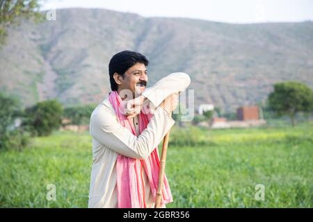Felice agricoltore indiano che tiene in mano bastone di legno in piedi in campo agricolo indossando abito tradizionale kurta, sorridente uomo con baffi e capelli neri
