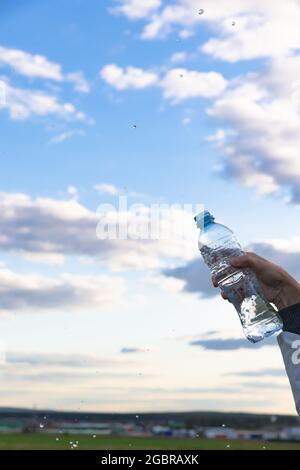 La mano di una donna solleva una bottiglia di acqua minerale pura altamente trasparente. Gli schizzi volano sullo sfondo di un cielo blu con nuvole bianche. S Foto Stock