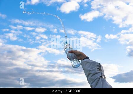 La mano di una donna solleva una bottiglia di acqua minerale pura altamente trasparente. Gli schizzi volano sullo sfondo di un cielo blu con nuvole bianche. S Foto Stock