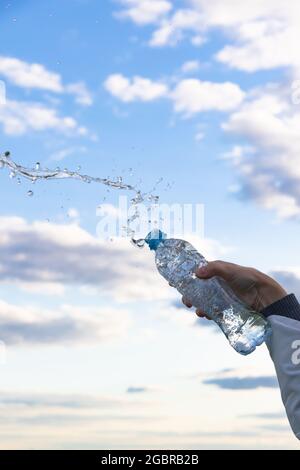 La mano di una donna solleva una bottiglia di acqua minerale pura altamente trasparente. Gli schizzi volano sullo sfondo di un cielo blu con nuvole bianche. S Foto Stock