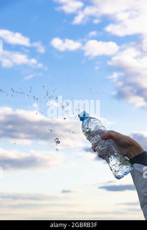 La mano di una donna solleva una bottiglia di acqua minerale pura altamente trasparente. Gli schizzi volano sullo sfondo di un cielo blu con nuvole bianche. S Foto Stock