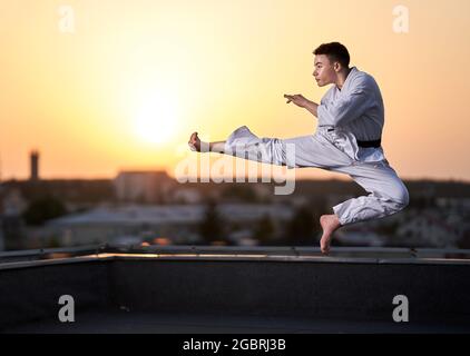 Giovane ragazzo praticante di karate in bianco kimono allenamento sul tetto al tramonto Foto Stock