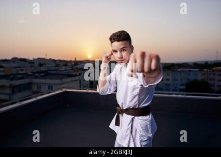 Giovane ragazzo praticante di karate in bianco kimono allenamento sul tetto al tramonto Foto Stock