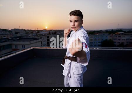 Giovane ragazzo praticante di karate in bianco kimono allenamento sul tetto al tramonto Foto Stock