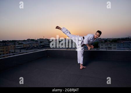 Giovane ragazzo praticante di karate in bianco kimono allenamento sul tetto al tramonto Foto Stock