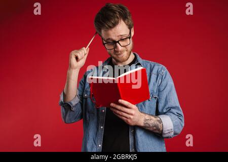 Pensivo giovane uomo ponderato in abiti casual in piedi su sfondo rosso parete, la lettura di un libro, tenendo la matita Foto Stock
