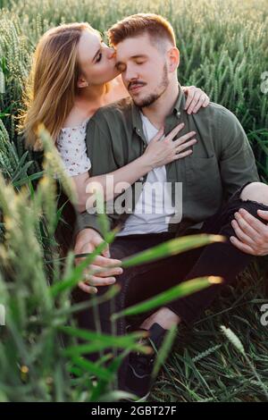 Bella giovane donna bacia suo marito la sera d'estate seduta in campo tra erba alta . Amore e tenerezza. Bei momenti di vita. Gioventù A. Foto Stock