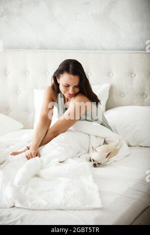 Concetto di mattina pigra. Bella donna felice si sveglia a letto e striscia le mani. Bella donna che riposa a casa in camera da letto. Il cane piccolo dorme vicino Foto Stock