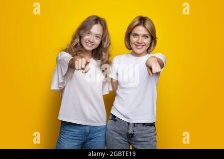 Le sorelle con i capelli biondi puntano verso la telecamera sorridente su una parete gialla dello studio che indossa t-shirt e jeans Foto Stock