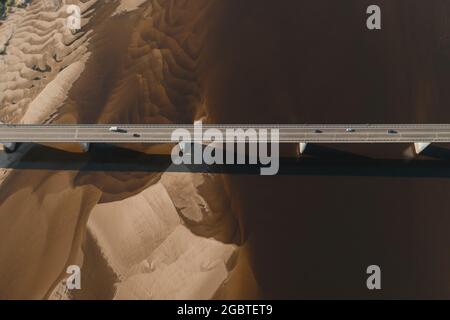 Un drone aereo ha girato delle automobili che guidano sul ponte sopra il fiume e sul lungofiume sabbioso al tramonto. Splendida vista dall'alto con una splendida costa con dune sabbiose. Ponte aereo drone con traffico auto Foto Stock
