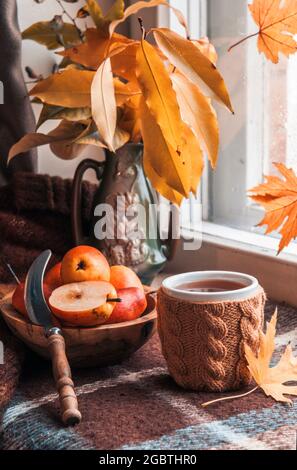 Tazza di tè nero caldo in una cassa di maglia beige, ciotola di legno con pere, foglie di autunno vicino alla finestra Foto Stock