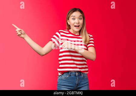 Ehi, sbrigati. Entusiasta eccitata cute asiatica bionda ragazza urbana puntando a sinistra mostrando offerte incredibili, aumentare sopracciglia entusiasta parlare fotocamera Foto Stock