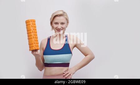 Ragazza che tiene un rullo di schiuma in mano sorridente. Ragazza carina pronta per un allenamento con reggiseno sportivo. Isolato su sfondo bianco. Fascia a rulli. Concetto di forma fisica e stile di vita sano. Foto Stock