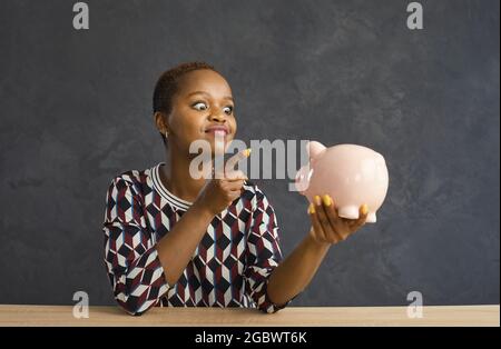 Divertente donna afroamericana punendo ondeggiando il dito alla banca piggy in mano. Foto Stock