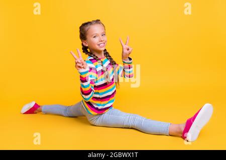 Foto di bambina fare spago mostra due v-segni usura camicia a righe jeans sneakers isolato colore giallo sfondo Foto Stock