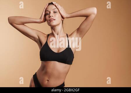 Giovane donna in costume da bagno che tiene la testa mentre si guarda fotocamera isolato sfondo beige Foto Stock