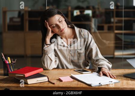 Giovane stanca donna bianca brunetta casual seduta dormendo alla scrivania dell'ufficio con gli occhi degli appunti chiusi Foto Stock