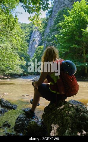 Ragazza con casco da arrampicata e uno zaino si siede su una roccia a Varghis gola, Persani montagne, Romania, vicino al fiume e pareti verticali roccia. Estate A. Foto Stock