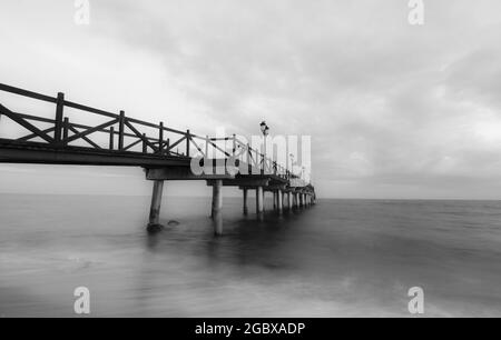 Immagine in scala di grigi di un molo sul mare con lunga esposizione sotto un cielo nuvoloso Foto Stock