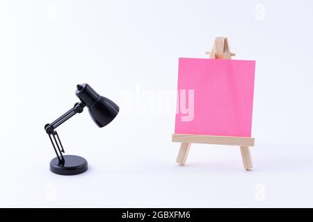 Easel in legno in miniatura con tela quadrata colorata vuota o carta Memo - Mockup. Mini supporto in legno con tavola da disegno pulita e piccola lampada da tavolo nera su sfondo bianco, Copy Space Foto Stock