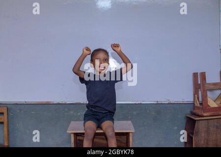 Bambina africana, pupilla o studente seduto di fronte a una lavagna bianca e con gioia spalancando le mani a causa dell'eccellenza nell'istruzione Foto Stock