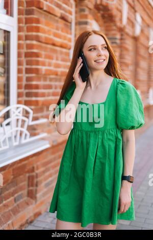 Verticale medio scatto ritratto di felice giovane donna che parla sul telefono cellulare che cammina sulla strada della città nel giorno d'estate. Foto Stock