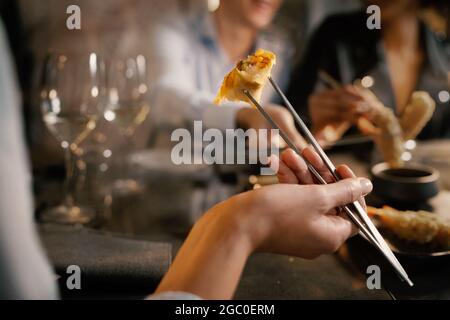 Dettaglio della mano di una donna che afferra un rotolo a molla con bacchette metalliche. Si siede a un tavolo con gli amici in un elegante ristorante di fusion food asiatico. Foto Stock