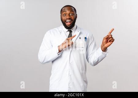 Eccitato, felice e sorpreso medico afro-americano che dice ai colleghi grandi notizie, indicando dita upprt angolo destro, sorridendo allegro e Foto Stock