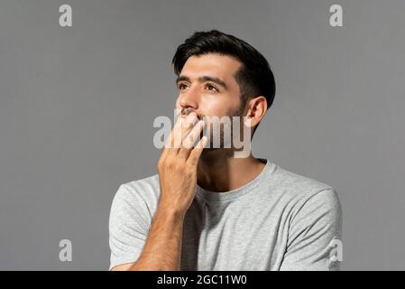 Scioccato giovane uomo caucasico guardando in su e pensando con la bocca di copertura della mano su sfondo grigio chiaro studio Foto Stock