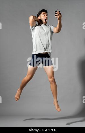 Ritratto a lunghezza intera di giovane uomo caucasico scioccato in sleepwear svegliarsi tardi guardando sveglia e saltando in uno sfondo grigio isolato studio Foto Stock