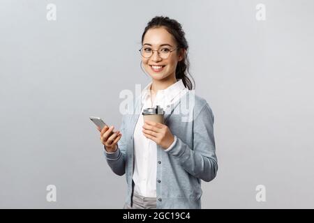 Stile di vita dell'ufficio, concetto di affari e di gente. Allegra ragazza asiatica attraente in bicchieri, tenendo take-away caffè e smartphone, guardare contento Foto Stock