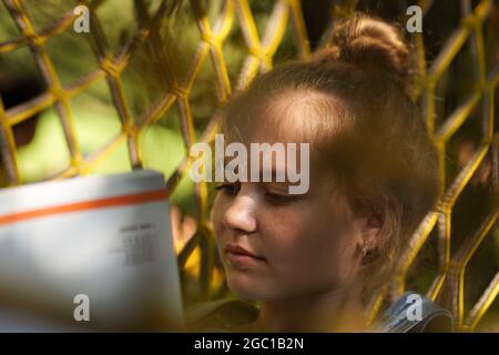 Giovane donna bionda studentessa sorridente leggendo UN libro mentre si steso su un amaca in giardino. Donna che si rilassa in amaca. Torna all'università scolastica Foto Stock