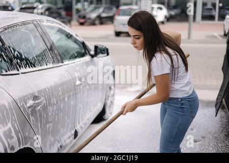 Attraente giovane donna che lava la sua auto con shampoo e spazzole. La donna lava l'automobile con schiuma e acqua all'esterno su autolavaggio self service Foto Stock
