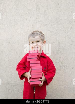 Concetto di consegna. Sorridente simpatico ragazzo di 4 - 5 anni in una divisa rossa tiene molte piccole scatole nelle sue mani. Consegna pacchi, piccolo postino, positivo, jo Foto Stock