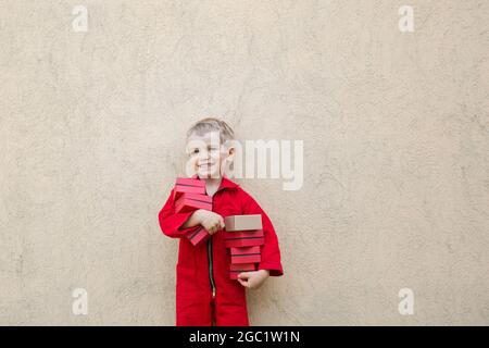 Sorridente simpatico bambino in una tuta rossa mantiene molte piccole scatole nelle mani. Consegna pacchi, piccolo postino, assistente. Positivo, gioia, gam Foto Stock