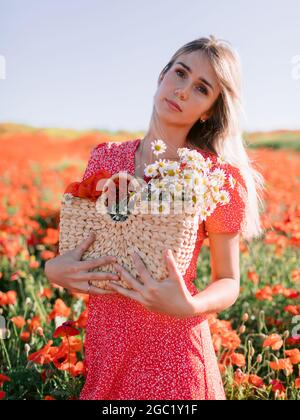 Attraente donna caucasica in un campo di papavero al tramonto. Concetto di libertà con colori tenui. Donna in abito rosso e borsa in vimini con fiori Foto Stock